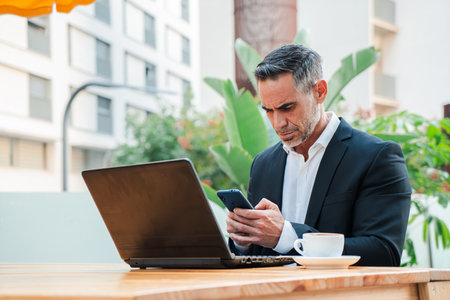 Focused businessman in a suit using a laptop and smartphone while working remotely at a terrace cafeの写真素材