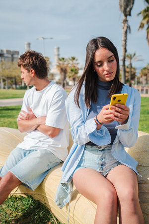 Young couple experiencing tension while one browses their smartphone in the sunny outdoor park settingの写真素材