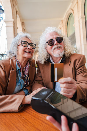 Joyful senior couple happily making a payment with a credit card while smiling and enjoying their time together at a charming cafe table in aの写真素材
