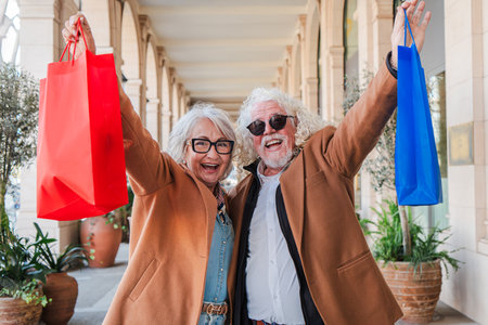 Joyful senior couple happily raising colorful shopping bags while enjoying a fun day out together in a lively shopping area filled with excitement andの写真素材