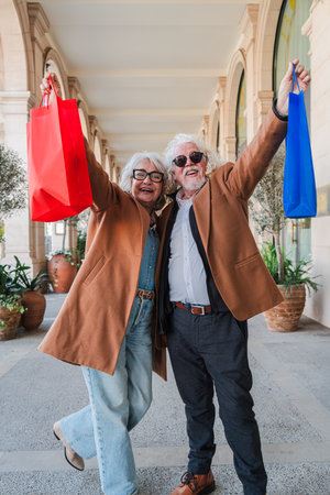 Joyful senior couple celebrating a successful shopping day, raising colorful bags with excitement while strolling together in a lively shoppingの写真素材
