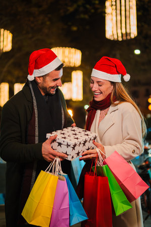 Vertical. Boyfriend giving a Christmas gift to his girlfriend outdoors at night, both wearing Santa hats and smiling with joy, surrounded by festiveの写真素材