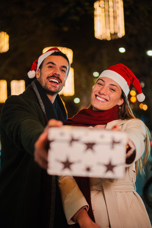 Vertical. Joyful Couple Wearing xmas Santa Hats Happily Holding a Gift Wrapped Box, Radiating...