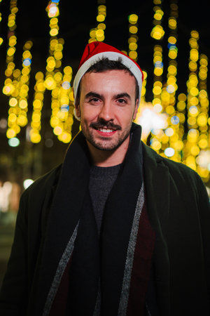 Vertical. Smiling young man wearing a Santa hat at night with festive glowing lights in the background, celebrating Christmas outdoors in an urbanの写真素材