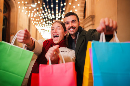 Happy couple holding colorful shopping bags under xmas festive lights, celebrating Christmas shopping and holiday spirit in an illuminated urban mallの写真素材