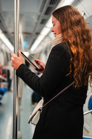 Enthusiastic young woman engaged in reading on her tablet while commuting on the subway during a busy day, showcasing a moment of focus and leisureの写真素材