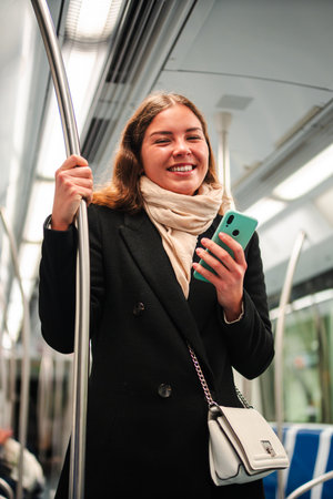 Young woman happily using her smartphone while standing on a subway train, engaging with applications and enjoying her commute in a lively and dynamicの写真素材
