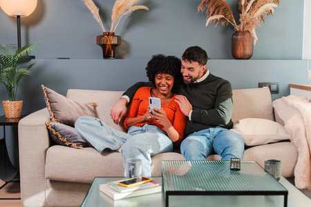 Joyful couple enjoying quality time together while using a smartphone on a cozy couch in their homeの写真素材