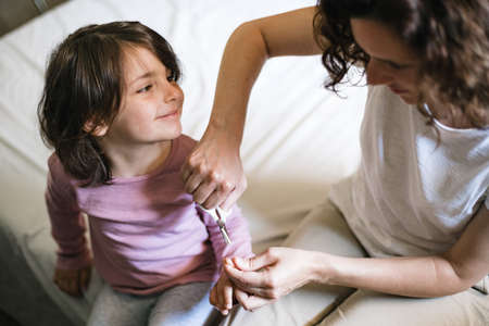 Young mother sitting on a bed carefully cuts her daughter's finger nails with scissors as she looks back with loveの写真素材