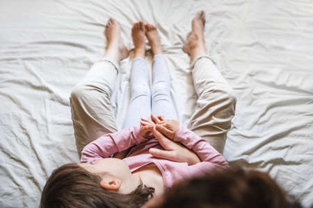 A little girl lays back on her mother's chest in the bed as they hold hands together. Mom wraps her in her arms as a sign of loveの写真素材