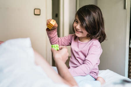 A little girl sitting on a bed plays with toy bricks with her motherの写真素材