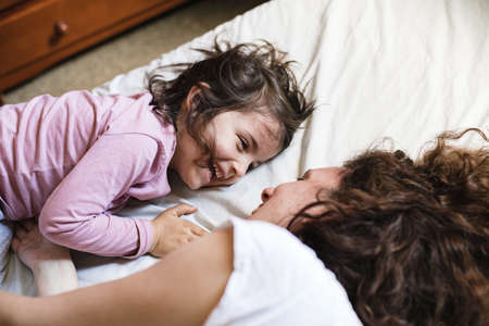A little girl lying down in her mother's bed laughs while they have a great time playing aroundの写真素材