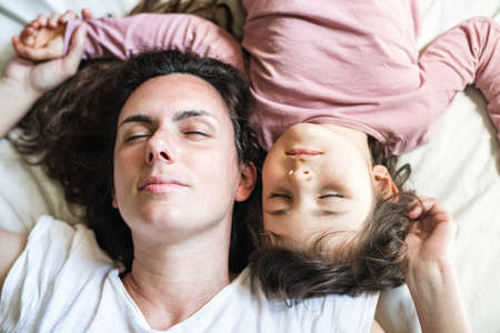 Mother and daughter lie down in bed with their heads facing up. Their eyes are closed having a...