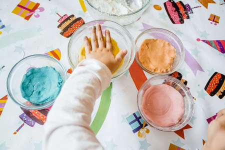 Little girl kneads colored dough to make homemade plasticine. She has many bowls with different colors sitting on a colorful party tableclothの写真素材