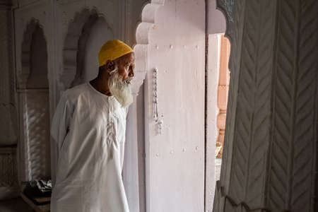 An old muslim man wearing a yellow beanie looks out the door in a mosque in Panchakki. He wears a yellow kufi and a white dress. He has growned a long white beard. Aurangabad, Indiaのeditorial素材