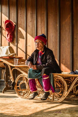 The old woman from the lisu hilltribe waits for some food sitting on a wicker bench. She wears a beanie, a jacket and a lisu traditional dress. Her feet hang in the air. The hut is made out of wood, cane and clay. Soppong village, Thailandのeditorial素材