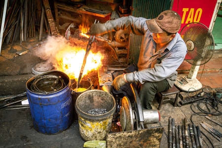 Fire bursts as a blacksmith plunges a piece of metal in chemicals and cools it down with water. Forge in the streets of the old quarter in Hanoi, Vietnamのeditorial素材