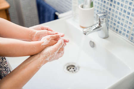 Mother and girl child washing hands together in bathroom sink with lots of soap under running water tap. Holding handsの写真素材