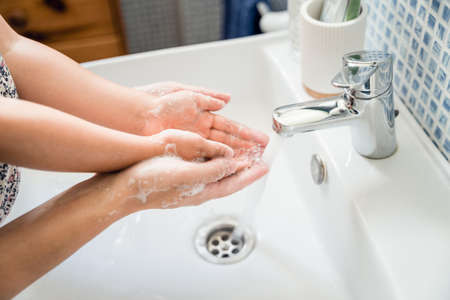 Mother and girl child washing hands together in bathroom sink with soap under running water tap to rinse off. Fingers hands extended open. High point of viewの写真素材