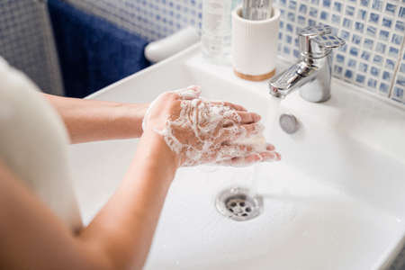 Young woman washes hands in bathroom washbasin under running tap water. Rubs hands together with soapの写真素材