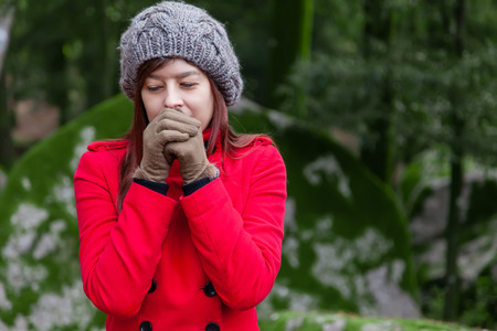 Young woman shivering with cold and blowing hot air to the hands on a forest wearing a red overcoat, a beanie and gloves during winterの写真素材