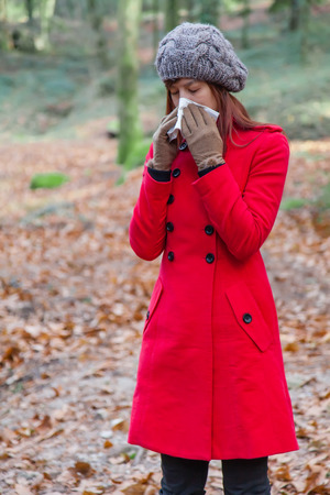 Young woman suffering from a cold or flu blowing her nose on a white paper handkerchief on a forest wearing a red overcoat, a beanie and gloves during winterの写真素材