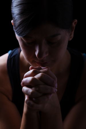 Low key portrait of a faithful woman praying, hands folded in worship to god, head down and eyes closed in religious fervor, on a black background. Concept for religion, faith, prayer and spiritualityの写真素材
