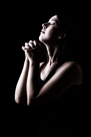 Faithful woman praying, hands folded in worship to god with head up and closed eyes in religious fervor, on a black background. Concept for religion, faith, prayer and spirituality.の写真素材