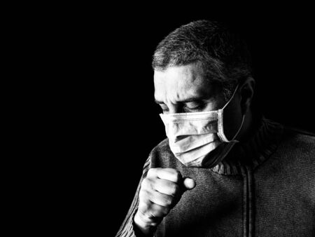 man with surgical mask sneezing or coughing. Pandemic or epidemic and scary, fear or danger concept. Protection for biohazard like COVID-19 aka Coronavirus. Black Background. Black and Whiteの写真素材