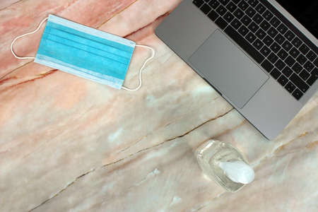 Top view of a computer accompanied by a mask and hydroalcoholic gel on a pink marble backgroundの写真素材