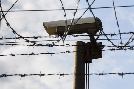 Security camera behind barbed wire fence stretched around prison walls