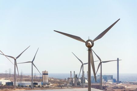 White turbines at wind farm with power station and sea in background, arid landscape on sunny summer day, sustainable renewable electricity conceptの写真素材
