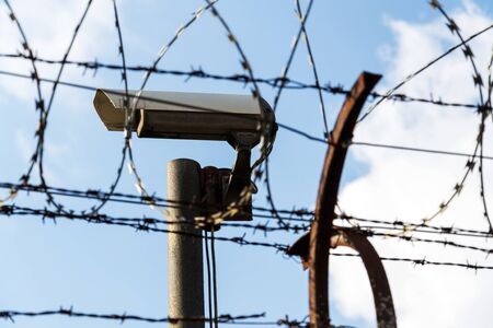 Security camera behind barbed wire fence on the wall, prison, security, crime or illegal immigration concept, blue sky backgroundの写真素材