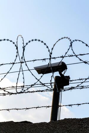 Security camera behind barbed wire fence on the wall, prison, security, crime or illegal immigration concept, blue sky backgroundの写真素材