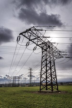 Sun hidden behind dramatic clouds and electricity pylons conducting current from distribution power station, solar renewable energy and grid stability concept, copy spaceの写真素材