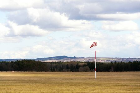 Red and white windsock on empty grassy airfield, dramatic clouds in background, windy weather forecast and air traffic conceptの写真素材