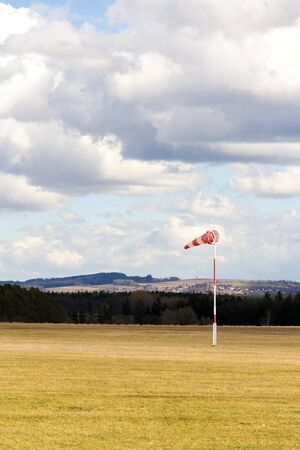 Red and white windsock on empty grassy airfield, dramatic clouds in background, windy weather forecast and air traffic conceptの写真素材
