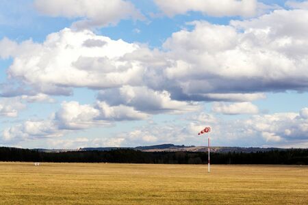 Red and white windsock on empty grassy airfield, dramatic clouds in background, windy weather forecast and air traffic conceptの写真素材