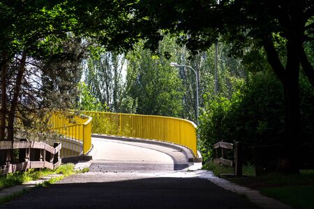 Yellow pedestrian bridge over road turning left from shadow of deciduous trees in park on sunny summer dayの写真素材