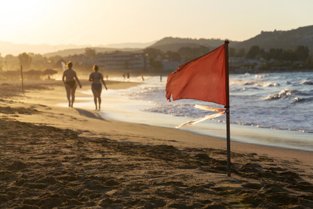 Unrecognizable people walking on a beach with a red flag warning of big waves at dawnの写真素材