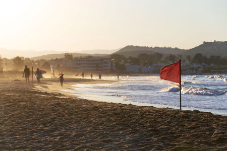 Unrecognizable people walking on a beach with a red flag warning of big waves at dawnの写真素材