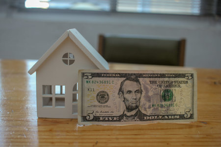 A conceptual shot featuring a small wooden house model and a five-dollar banknote. Represents the decision between keeping money in liquid cash or investing in property and real estate assets.の写真素材