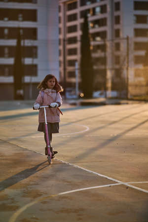 girl in coat and pink scooter raising her leg and smiling while driving during sunsetの写真素材