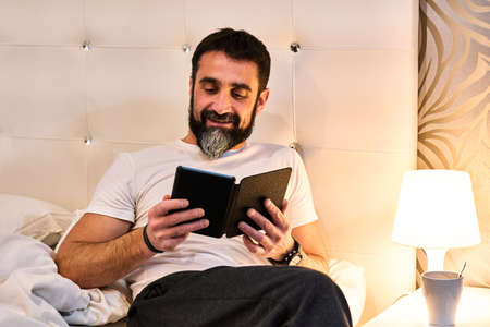 bearded man reading an e-book in bed with a cup of coffee or infusion on the coffee table