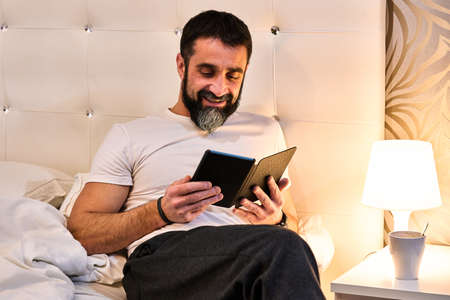 bearded man reading an e-book in bed with a cup of coffee or infusion on the coffee table