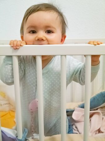 Cute little baby. She is standing in her cradle, chewing it.の写真素材