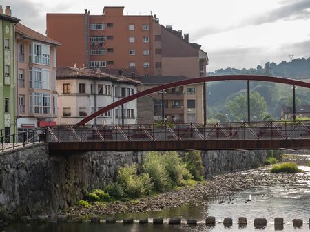 Metallic bridge crossing a quiet river. You can see some houses next to it.の写真素材