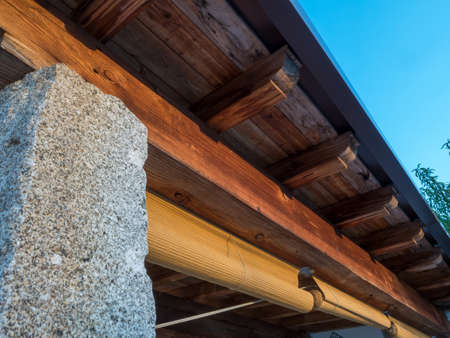 Rustic porch with wooden beams, stone columns and blinds, with a blue sky and some trees in the...