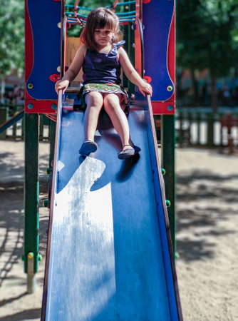 A girl in a colorful dress playing on a playground, going down a slide on a sunny summer day