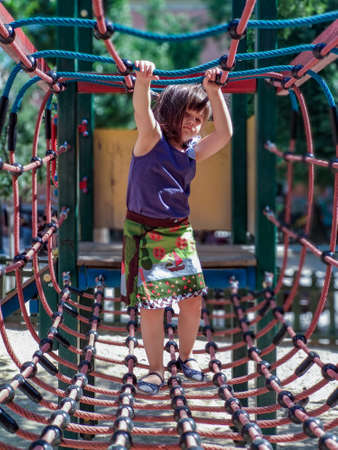 Girl in a colorful dress on a playground while playing across a rope bridge on a sunny summer dayの写真素材